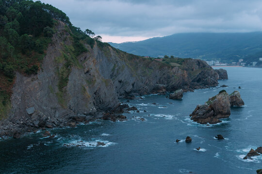 Rugged cliff and rocky shore jutting into dark blue atlantic, green vegetation clinging to textured flysch formations under moody cloudy sky &mdash; dramatic, remote coastal landscape
