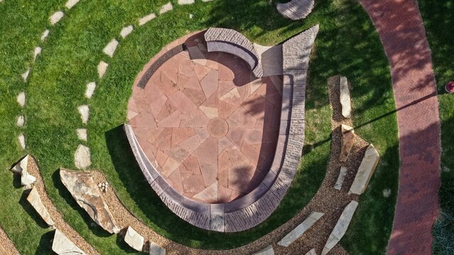 Aerial view of a circular stone plaza with curved seating benches surrounded by lush green grass and a winding brick pathway in Denver, Colorado, United States.