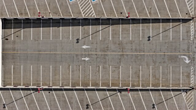 Aerial view of an empty parking lot with white painted parking spaces, directional arrows, and long shadows cast by light poles in Denver, Colorado, United States.