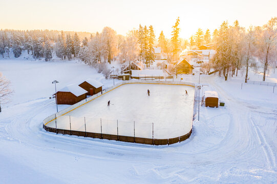 Aerial view of an outdoor ice rink with people playing hockey surrounded by snow-covered trees and wooden cabins during golden hour in Latvia.