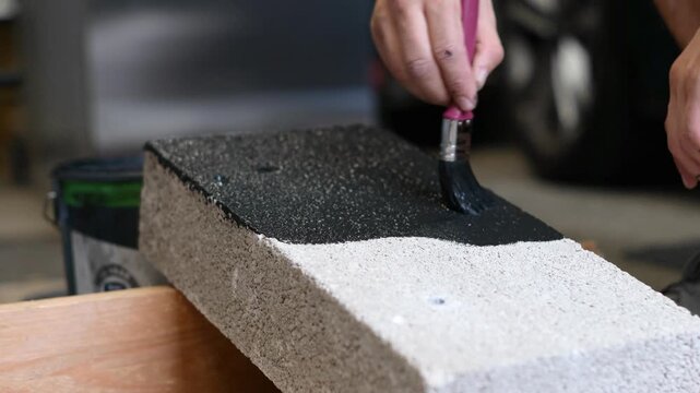Close-up view of a man carefully covering the rough top surface of a concrete masonry unit with dark paint