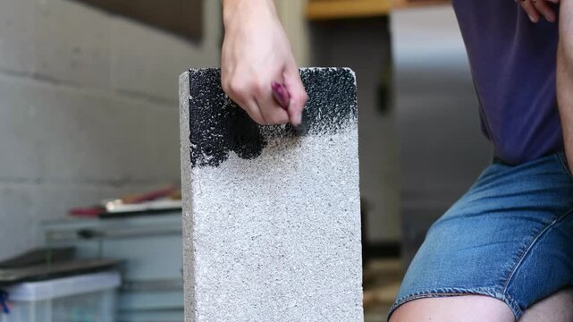 Close-up of a man applying black paint to the rough surface of a concrete masonry block during a construction or renovation project