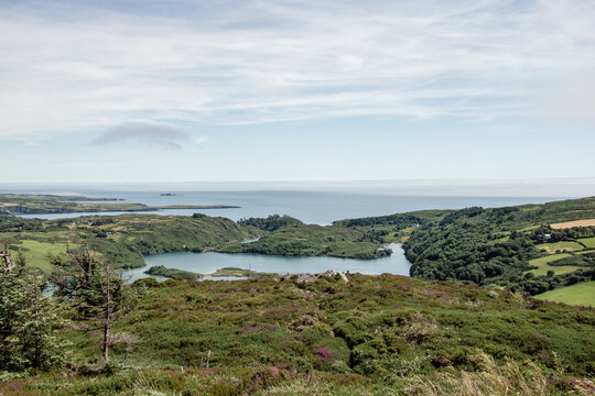Scenic aerial view of a green coastal landscape with a calm bay, rolling hills covered in heather, and the ocean stretching into the horizon under a cloudy sky