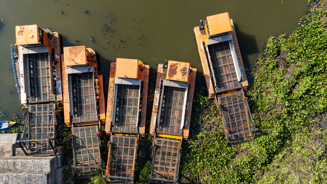 Aerial view of orange aquatic weed harvester machines docked along a riverbank with lush green vegetation in Salatiga, Central Java, Indonesia.