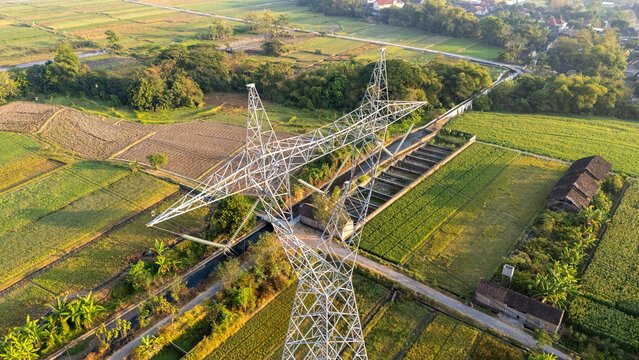 Aerial view of a high-voltage electricity pylon standing amidst lush green rice fields and irrigation channels in the rural landscape of Klaten, Central Java, Indonesia.