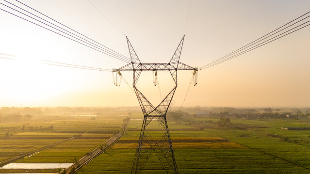 Aerial view of a high-voltage electricity pylon standing amidst vast green rice fields during the golden hour in Klaten, Central Java, Indonesia.