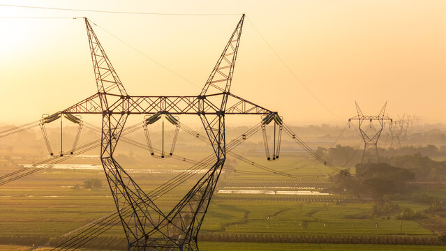 Aerial view of a high-voltage electricity transmission tower standing over rural rice fields during the golden hour in Klaten, Central Java, Indonesia.