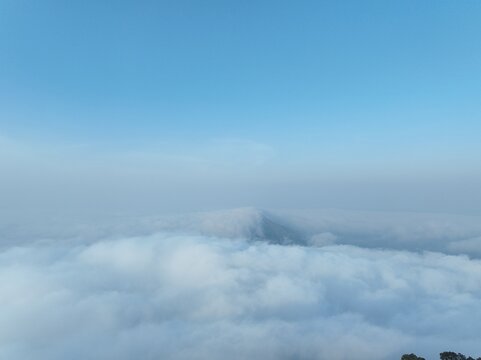 Aerial view of Triangle Sky Telomoyo peak rising above a dense sea of clouds under a clear blue sky in Kecamatan Ngablak, Jawa Tengah, Indonesia.