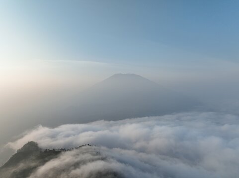Aerial view of the misty peak of Triangle Sky Telomoyo rising above a dense sea of clouds in the early morning light in Kecamatan Ngablak, Jawa Tengah, Indonesia.