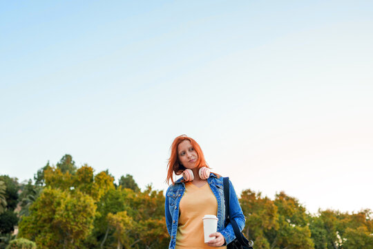 Young red haired woman holding coffee cup and wearing headphones outdoors, contemplating a casual moment