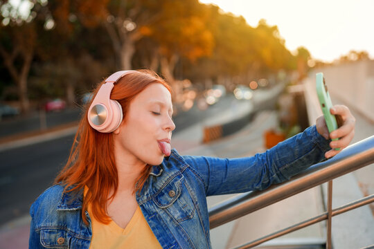 Young woman sticking out tongue for a playful selfie using a smartphone and pink headphones outdoors