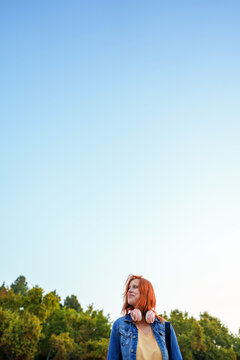 Young woman with red hair wearing headphones around neck looking away outdoors on a clear day