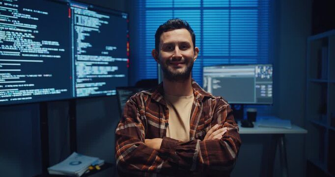 Portrait of confident Caucasian male programmer smiles looking at camera in office at night. Professional developer workspace, Secure computer network system, Advanced cybersecurity data concept.