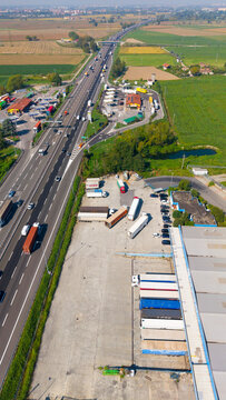 Aerial view of the A50 Tangenziale Ovest di Milano highway with heavy traffic, a service station, and a large logistics depot with parked semi-trucks and trailers in Milano, Lombardia, Italy.