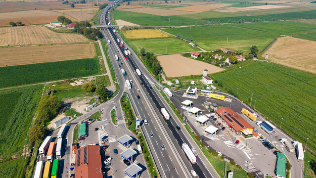 Aerial view of A50 Tangenziale Ovest di Milano highway with heavy traffic, service stations on both sides, surrounded by agricultural fields and green farmland in Milano, Lombardia, Italy.