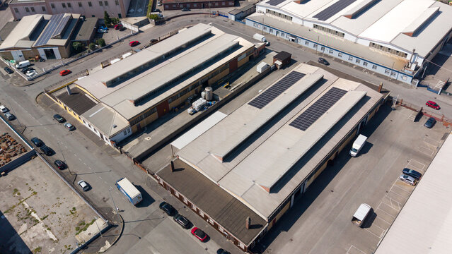 Aerial view of industrial warehouses with solar panels on rooftops, parked cars, and trucks in an industrial zone in Sesto Ulteriano, Lombardy, Italy.