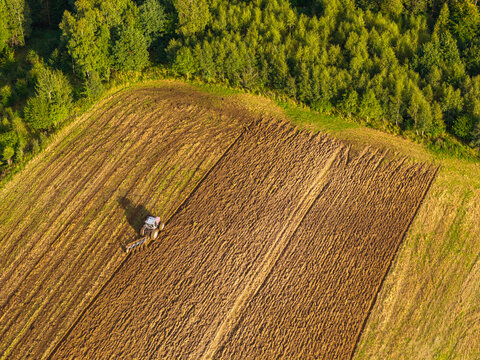 Aerial view of a tractor plowing a field with deep furrows in the soil, bordered by a lush green forest in the Kashubia region, Pomeranian Voivodeship, Poland.
