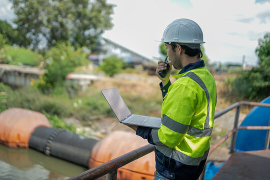 Male environmental engineer communicates by radio while using laptop during environmental monitoring and water infrastructure assessment at drainage facility.