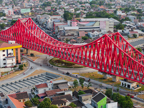 Aerial view of the vibrant red Jembatan Kereta Api Joglo railway bridge spanning over a busy roundabout and urban landscape in Surakarta, Jawa Tengah, Indonesia.