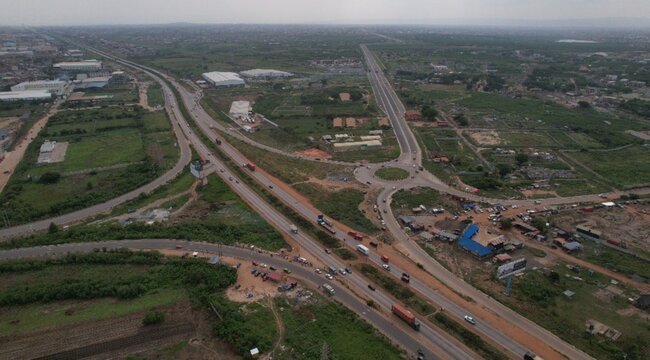 Aerial view of a highway intersection with a roundabout, trucks and cars driving on the road, surrounded by green fields and industrial buildings in Ghana.