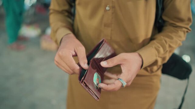 Medium close up video shot of a man wearing a brown kurta holding and opening an empty leather wallet outdoors on a city street.