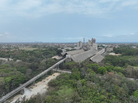 Aerial view of the industrial cement plant facility with its extensive conveyor belt system and large factory buildings surrounded by lush green trees in Cilacap, Central Java, Indonesia.