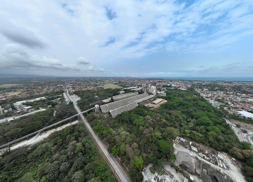 Aerial view of the industrial cement plant complex surrounded by lush forest, with the urban cityscape and coastline visible in the distance in Cilacap, Central Java, Indonesia.