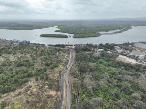 Aerial view of a road leading towards the river estuary surrounded by lush mangrove forests and industrial facilities in the coastal landscape of Cilacap, Central Java, Indonesia.
