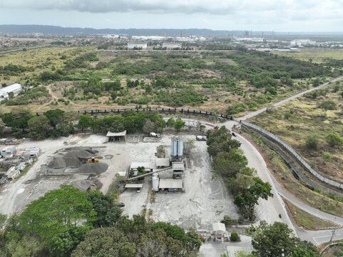 Aerial view of an industrial concrete mixing plant with silos, gravel piles, and heavy machinery, surrounded by trees and open land in Cilacap, Central Java, Indonesia.