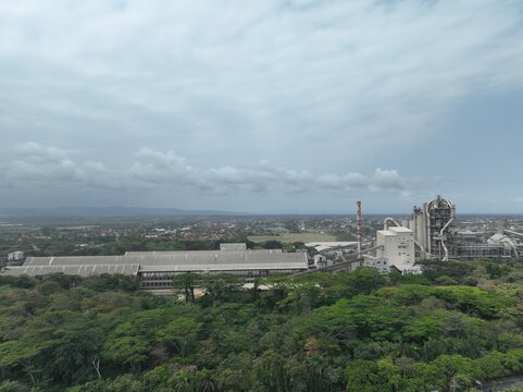 Aerial view of a large industrial cement plant complex surrounded by lush green trees under a cloudy sky in Cilacap, Central Java, Indonesia.