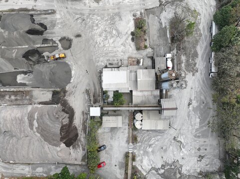 Aerial view of an industrial concrete processing plant with large piles of sand and gravel, a yellow loader, storage silos, and trucks in Cilacap, Central Java, Indonesia.