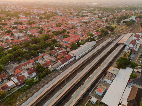 Aerial view of Stasiun Kejaksan railway station with trains on tracks, surrounded by dense urban rooftops and lush trees in Cirebon, West Java, Indonesia.