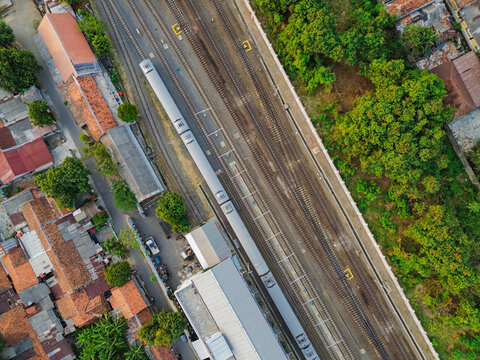 Aerial view of Stasiun Kejaksan featuring a passenger train on the tracks, surrounding urban rooftops, and lush green trees in Cirebon, West Java, Indonesia.