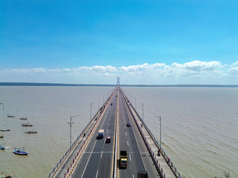 Aerial view of Suramadu Bridge spanning the Madura Strait with traffic on the road and small fishing boats in the water under a clear blue sky in Surabaya, East Java, Indonesia.