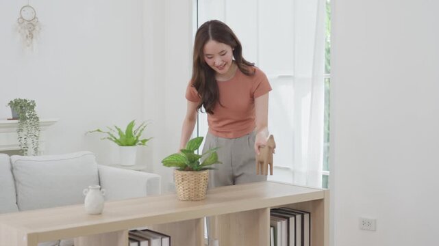 Asian adult woman cleaning wooden console table in bright living room with feather duster and potted plant, maintaining modern home interior near sunlit window during calm household chore scene
