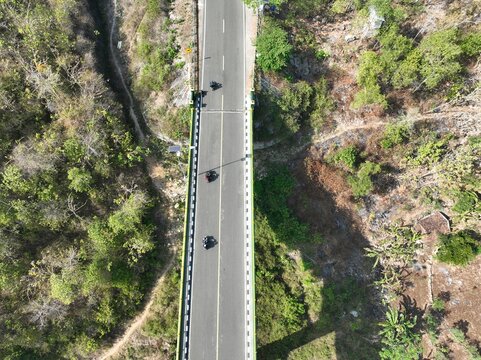 Aerial view of a road bridge surrounded by lush forest with motorcycles traveling along the asphalt surface in Gunungkidul Regency, Special Region of Yogyakarta, Indonesia.