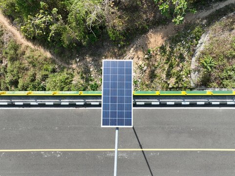 Aerial view of a solar panel on a pole beside a paved road with yellow lane markings, surrounded by vegetation in Gunungkidul Regency, Special Region of Yogyakarta, Indonesia.