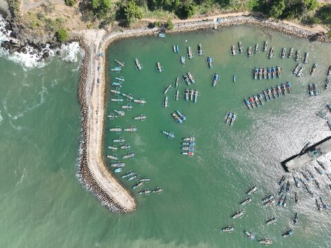 Aerial view of numerous traditional fishing boats anchored in the turquoise harbor waters protected by a stone breakwater along the coastline in Pacitan, East Java, Indonesia.