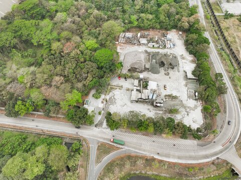 Aerial view of an industrial processing site with piles of gravel and sand, surrounded by dense green forest and adjacent to a winding road in Cilacap Regency, Central Java, Indonesia.