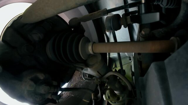 Drive shaft and CV joint moving with front suspension while car travels on road. Close underbody view of axle transferring rotation toward wheel hub. Steering linkage and control arm working beside