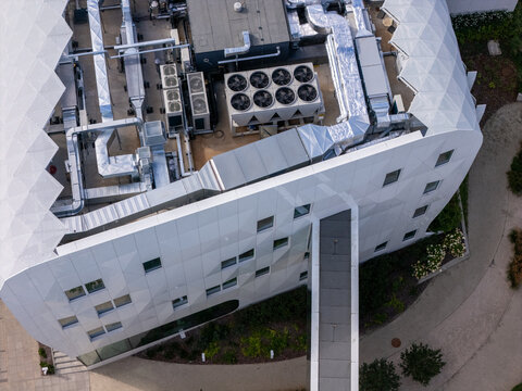 Aerial view of a modern medical complex featuring a geometric white facade, rooftop HVAC systems, and a connecting bridge over a landscaped path in Gdansk, Poland.