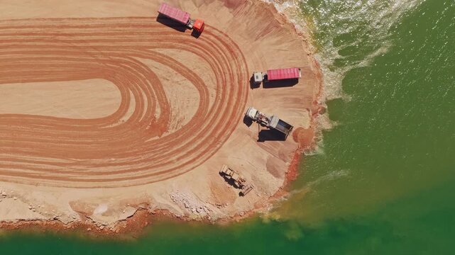 Top-down aerial view of heavy machinery operating at a construction site, with a dumper truck unloading sand into the water and a bulldozer leveling the ground near the green lake shoreline