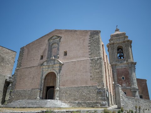 Majestic medieval church exterior in Erice, Italy, showcasing a towering stone campanile, classical arched doorway, and ancient masonry bathed in bright sunlight.