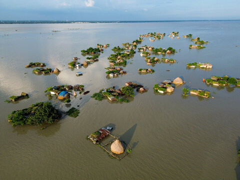 Aerial view of a flooded rural landscape with small islands of traditional huts and lush trees surrounded by expansive floodwaters in Rajshahi District, Rajshahi Division, Bangladesh.