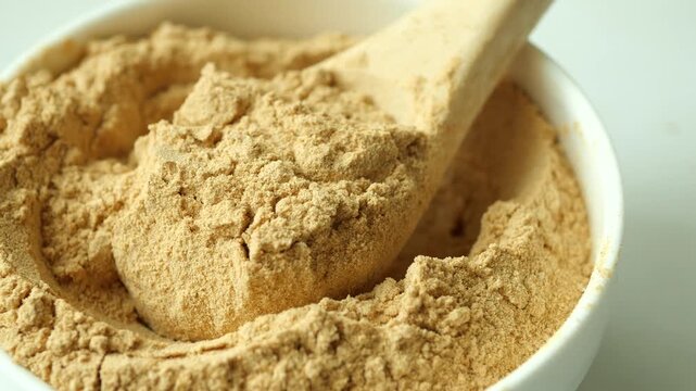 Close up slow motion video of wooden spoon filled with golden brown maca powder in white bowl on table