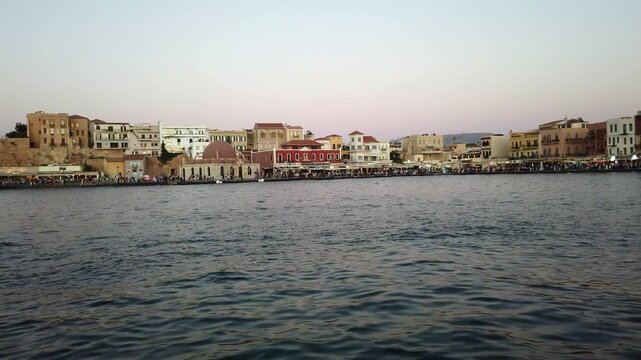 View from the harbor tower across choppy waters to the waterfront shops and cafes lining the Chania harbor front in Crete, Greece.