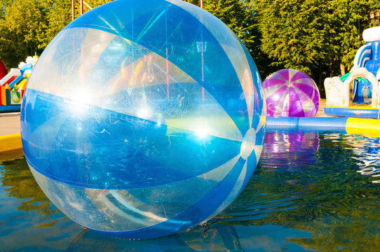Amusement park background. A large blue inflatable zorb ball floating on water. Green trees and colorful inflatable attractions in a sunny outdoor pool