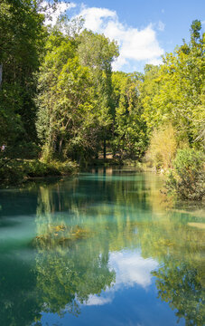 Vista vertical de un sereno r&iacute;o de aguas turquesas reflejando los &aacute;rboles del bosque y las nubes sobre un cielo azul brillante.