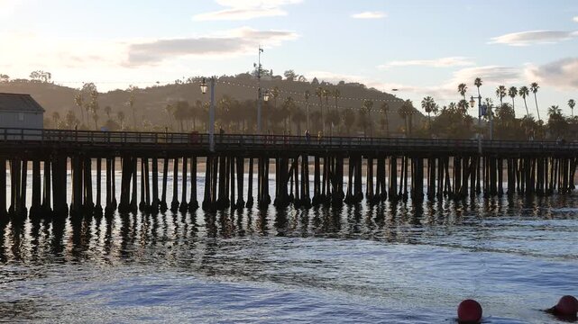 Sunset at Santa Barbara, California from Stearns Wharf, January 2024. Palm trees, recreationists and tourists walk the pier and enjoy the beautiful weather. With sun flare.