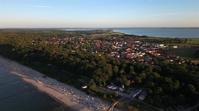 Red-roofed town spreads along tree lined coastline, curved pier extends across sandy beach crowded with visitors, warm sunset light bathes rooftops near Usedom, Germany. Aerial drone wide shot